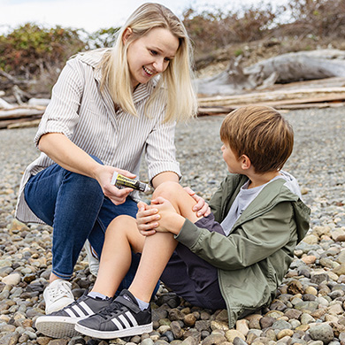 Young boy sitting on a pebble beach with his jacket on and his mother applying ointment on his knee while smiling.